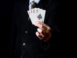 A man in a black suit and a tie holds 4 aces in his hand with black background. Poker cards. Play...