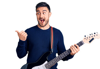Young handsome man playing electric guitar pointing thumb up to the side smiling happy with open mouth