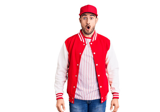 Young handsome man wearing baseball jacket and cap scared and amazed with open mouth for surprise, disbelief face