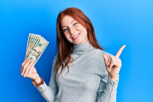 Young red head girl holding 20 dollars banknotes smiling happy pointing with hand and finger to the side
