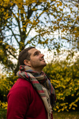 Photo of a young and attractive man wearing a scarf looking to the trees that surround him, enjoying a beautiful autumn day in nature