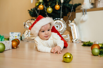 Cute smiling baby is lying under a festive Christmas tree and playing with gifts. Christmas and New Year celebrations