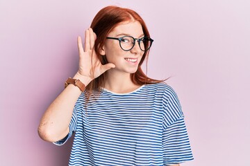 Young red head girl wearing casual clothes and glasses smiling with hand over ear listening and hearing to rumor or gossip. deafness concept.