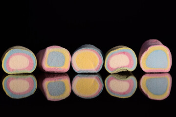 Several brightly colored marshmallows in pastel colors lie next to each other in front of a dark background and are reflected