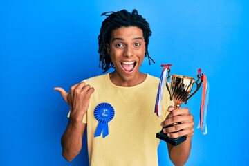 Young african american man wearing first place badge holding trophy pointing thumb up to the side smiling happy with open mouth