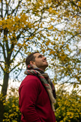 Photo of a young and attractive man wearing a scarf looking to the trees that surround him, enjoying a beautiful autumn day in nature