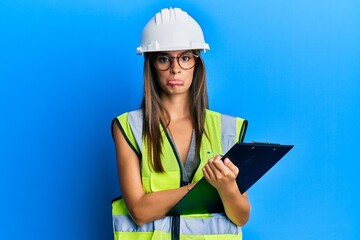 Young hispanic woman wearing safety helmet holding clipboard depressed and worry for distress, crying angry and afraid. sad expression.