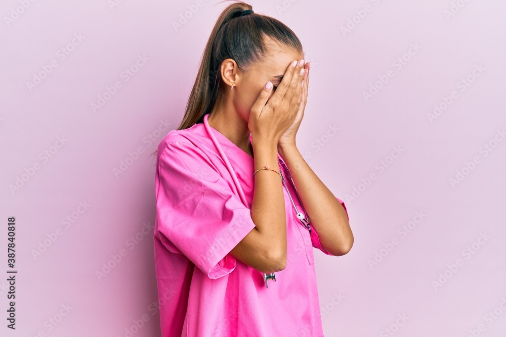 Wall mural Young hispanic woman wearing doctor uniform and stethoscope with sad expression covering face with hands while crying. depression concept.