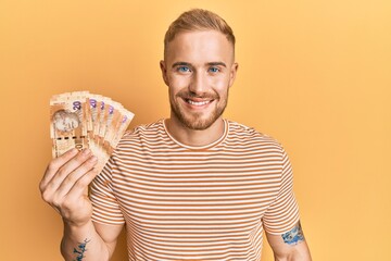 Young caucasian man holding south african 20 rand banknotes looking positive and happy standing and smiling with a confident smile showing teeth