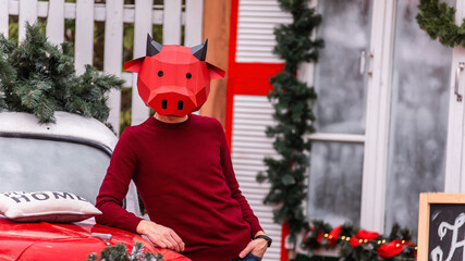 Obraz premium A young man in a bull ox costume is a symbol of the new year against the background of a red car, a festive Christmas tree, decorated with garlands of porch lights with a wreath. Copy space, template