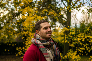 Photo of a young and attractive man wearing a scarf looking to the trees that surround him, enjoying a beautiful autumn day in nature