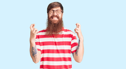 Handsome young red head man with long beard wearing casual clothes gesturing finger crossed smiling with hope and eyes closed. luck and superstitious concept.