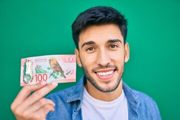 Young latin man smiling happy holding new zealand 100 dollar banknote at the city.