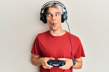 Young hispanic man playing video game holding controller making fish face with mouth and squinting eyes, crazy and comical.