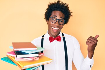 Handsome african american nerd man with afro hair holding books pointing thumb up to the side smiling happy with open mouth