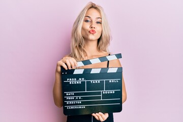 Beautiful caucasian blonde girl holding video film clapboard looking at the camera blowing a kiss...