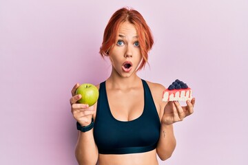 Young beautiful redhead woman holding green apple and cake slice afraid and shocked with surprise and amazed expression, fear and excited face.