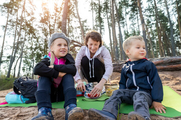 Young adult caucasian beautiful mother enjoy having fun eating snack on picnic with two cute little children in pine forest outdoors on warm autumn day. Family nature outdoor recreation lifestyle