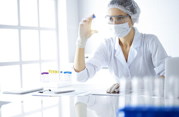 Professional female scientist in protective eyeglasses researching tube with reagents in sunny laboratory. Medicine and science researching