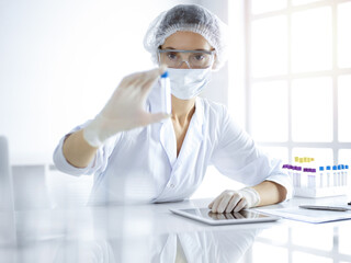 Professional female scientist in protective eyeglasses researching tube with reagents in sunny laboratory. Medicine and science researching