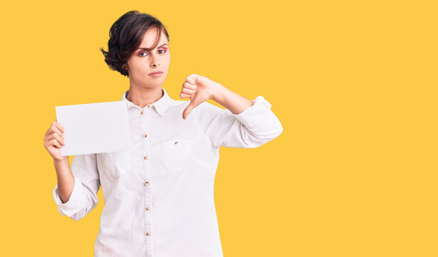 Beautiful young woman with short hair holding blank empty banner with angry face, negative sign showing dislike with thumbs down, rejection concept