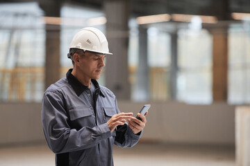 Side view portrait of mature worker using smartphone while standing at construction site or in...