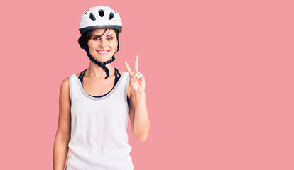 Beautiful young woman with short hair wearing bike helmet smiling with happy face winking at the camera doing victory sign. number two.