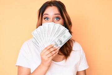 Young beautiful caucasian woman holding dollars covering mouth with hand, shocked and afraid for mistake. surprised expression