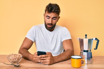 Young hispanic man sitting on the table having breakfast and using smartphone scared and amazed with open mouth for surprise, disbelief face