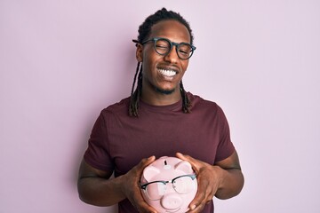 African american man with braids holding piggy bank with glasses winking looking at the camera with...