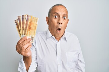 Handsome senior man holding 500 norwegian krone banknotes scared and amazed with open mouth for surprise, disbelief face