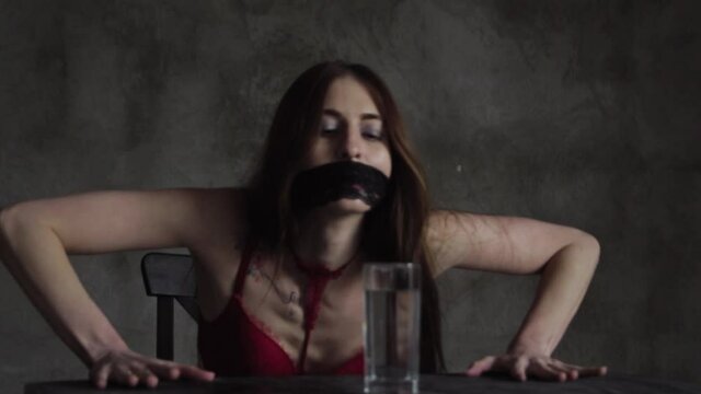 A girl in red underwear and gagged sits at a table in front of a glass of water. The woman is thirsty