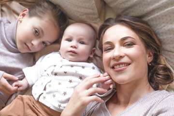 Mother holding infant baby, take care of her 3 months old child at home bedroom . Happy mom carying of her newborn son . Mother and elder sister kissing the boy