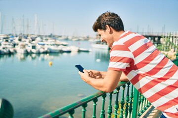 Young caucasian man smiling happy using smartphone leaning on the balustrade at the river.