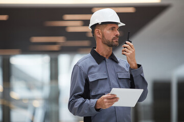 Waist up portrait of handsome mature worker speaking by walkie-talkie while supervising work at...