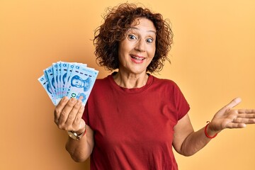 Beautiful middle age mature woman holding 10 yuan chinese banknotes celebrating achievement with happy smile and winner expression with raised hand