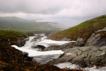 A bridge over a fast-flowing mountain stream, and snow on a Norwegian highland during a rainy and foggy summer day