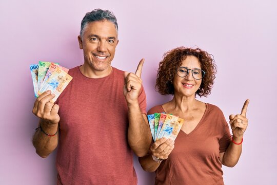 Beautiful middle age couple together holding swiss franc banknotes smiling happy pointing with hand and finger to the side
