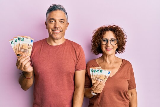 Beautiful middle age couple holding bunch of 50 euro banknotes looking positive and happy standing and smiling with a confident smile showing teeth