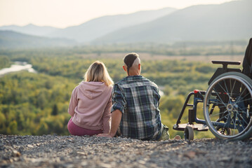 Pretty woman and her incapacitated husband resting together near his wheelchair on the hill.