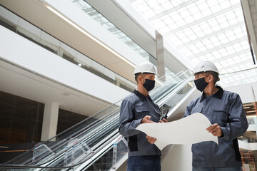 Low angle portrait of two construction workers wearing masks and discussing plans while standing in...