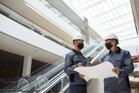 Low Angle Portrait Of Two Construction Workers Wearing Masks And Discussing Plans While Standing In Shopping Mall Or Office Building, Copy Space