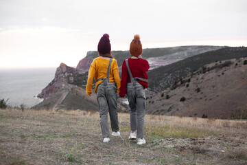 Back view of two preteen girls traveler wearing denim overalls, yellow and red sweater and knitted hats sit on top of the mountain landscape and looking to the sea.
