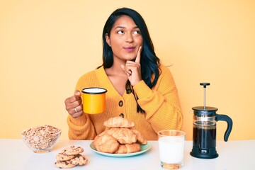 Beautiful latin young woman with long hair sitting on the table having breakfast serious face...