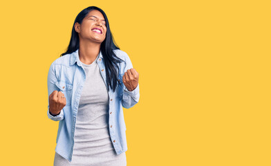 Hispanic woman with long hair wearing casual denim jacket very happy and excited doing winner gesture with arms raised, smiling and screaming for success. celebration concept.