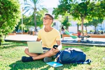 Young hispanic student smiling happy using laptop sitting on the grass at the university campus