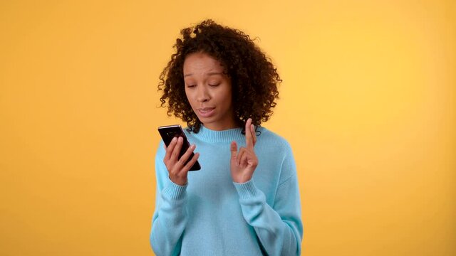 Good luck and winnings. young beautiful dark-skinned woman looks at the smartphone with great hope crossing her fingers. woman jumping for joy. in a blue sweater. person on isolated yellow background
