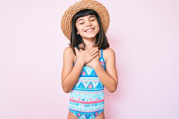 Young little girl with bang wearing swimsuit and summer hat smiling with hands on chest, eyes closed with grateful gesture on face. health concept.
