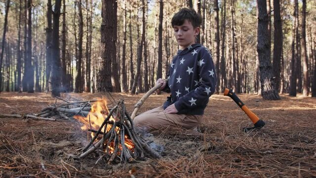 Happy boy tourist lights bonfire on family picnic in woods slow motion on the background of pine trees in summer. The ax is driven into the ground nearby. Family weekend. Hiking. Childhood. 