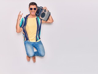 Young handsome hispanic man wearing sunglasses with open mouth and tongue mouth. Holding boombox doing horns sing jumping over isolated white background
